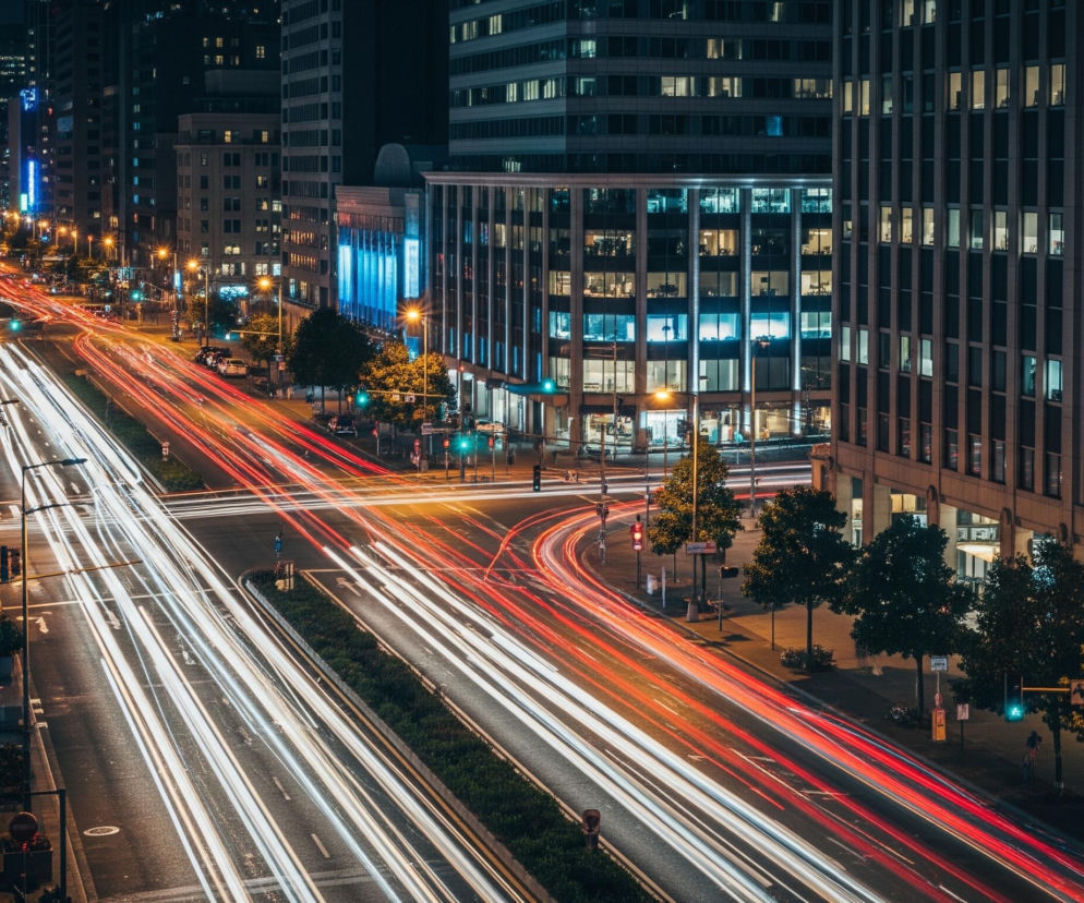 Ulica miejska oświetlona jest smugami czerwonych i białych świateł, tworząc wrażenie ruchu w nocy. Po obu stronach drogi wznoszą się wysokie budynki, z których wiele ma podświetlone okna w różnych odcieniach, od żółtego po niebieski. Na środku ulicy znajduje się zielony pas rozdzielający, a wzdłuż chodników rosną drzewa i stoją latarnie uliczne, które emitują ciepłe, pomarańczowe światło.