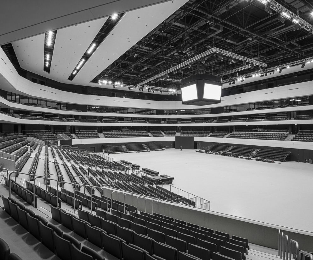 Wnętrze dużej hali sportowo-widowiskowej jest czarno-białe z niewielkimi, rozmytyymi kolorowymi elementami. Widoczne są puste rzędy siedzeń w ciemnym odcieniu, które otaczają centralne, jasne, prostokątne pole areny. Na środku areny po prawej stronie, w głębi obrazu, idzie pojedyncza, ciemna postać, a bliżej, po prawej stronie, rozmazana postać w czerwonej bluzce i ciemnych spodniach wchodzi po schodach, poruszając się w kierunku dołu obrazu.

Sufit hali jest konstrukcji otwartej z widocznymi kratownicami, reflektorami i systemami oświetlenia, z których część jest rozświetlona, tworząc jasne punkty. Z sufitu zwisa również duży, kwadratowy ekran z czterema jasnymi, pustymi panelami. Poziomy balkony o falistych, białych barierkach i ciemnych przeszkleniach opasują całą przestrzeń.