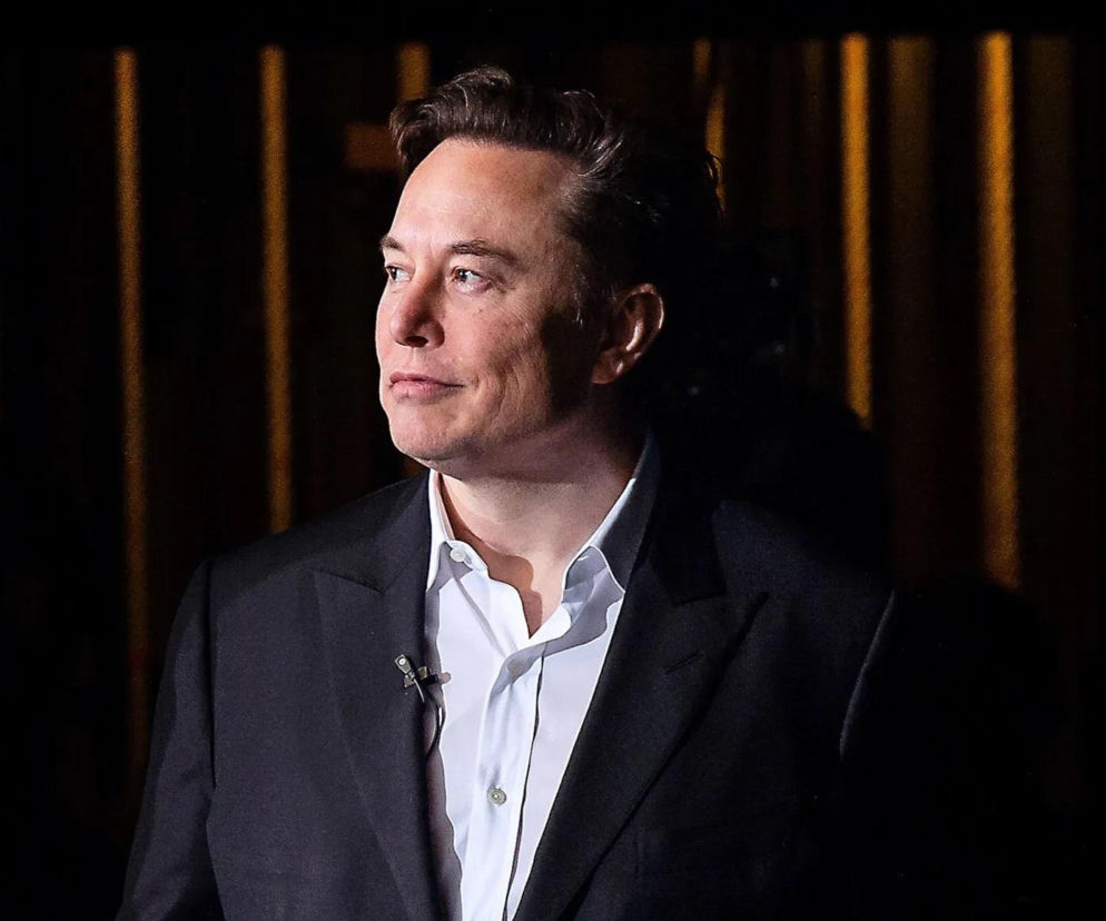 Elon Musk w ciemnym garniturze i białej koszuli patrzy w bok. O nowej polityce firmy Starlink przeczytasz w serwisie Eska.