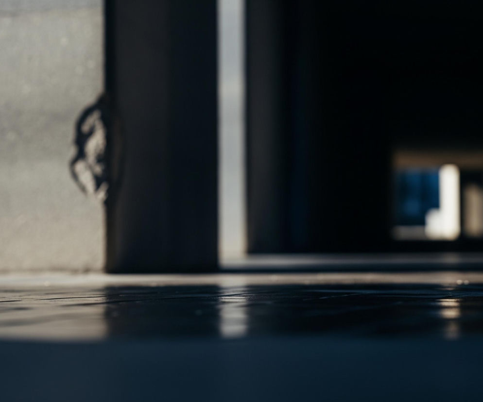 Ciemna, rozmyta powierzchnia wypełnia dolną część kadru, odbijając nieco jaśniejsze punkty światła z dalszej części sceny. Lewa strona obrazu przedstawia jasnoszarą, lekko chropowatą ścianę, na której widoczne są drobne nierówności i przebarwienia. Po prawej stronie tej ściany znajduje się ciemny, pionowy element, być może framuga lub krawędź otworu, a za nim rozciąga się ciemna przestrzeń z niewyraźnym, jaśniejszym prostokątem w oddali, sugerującym kolejne przejście lub okno.