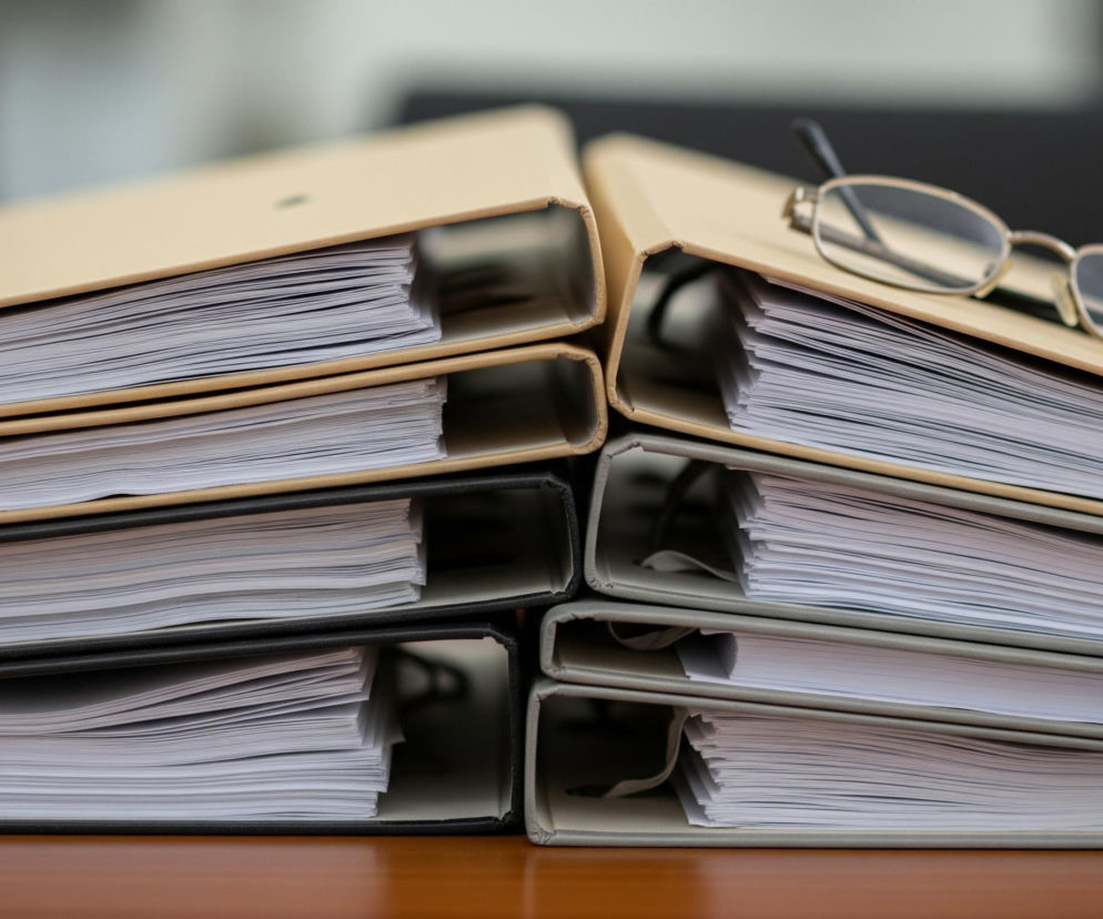 Sześć segregatorów z papierami leży ułożonych w dwa równoległe stosy na brązowej, błyszczącej powierzchni. Górny segregator w prawym stosie, o beżowej okładce, ma na sobie okulary ze złotą oprawką leżące skośnie. Cztery górne segregatory mają beżowe okładki, a dwa dolne są szare lub ciemnoszare, wszystkie wypełnione są białymi kartkami papieru. Tło jest rozmyte, z widocznymi ciemniejszymi i jaśniejszymi plamami.