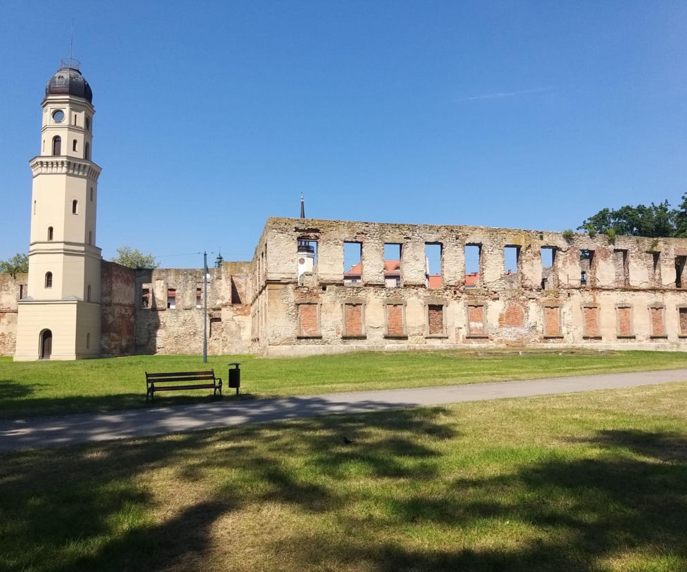 Zrujnowany zamek w Strzelcach Opolskich z odbudowaną wieżą i ruinami murów dawnego pałacu na tle błękitnego nieba. Na trawniku przed zamkiem ścieżka, ławka i kosz na śmieci. Przebudowa zamku na mieszkania, o czym można przeczytać na Architektura Murator Plus.