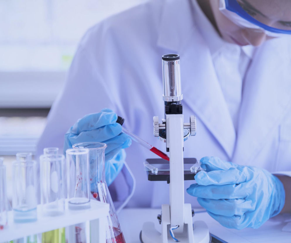 Naukowiec w białym fartuchu i niebieskich rękawiczkach laboratoryjnych, w goglach ochronnych, operuje mikroskopem, nanosząc próbkę czerwonej cieczy pipetą na szkiełko, obok probówki z płynami, co ilustruje postęp w badaniach nad szczepionkami, o czym przeczytasz na Poradnik Zdrowie.