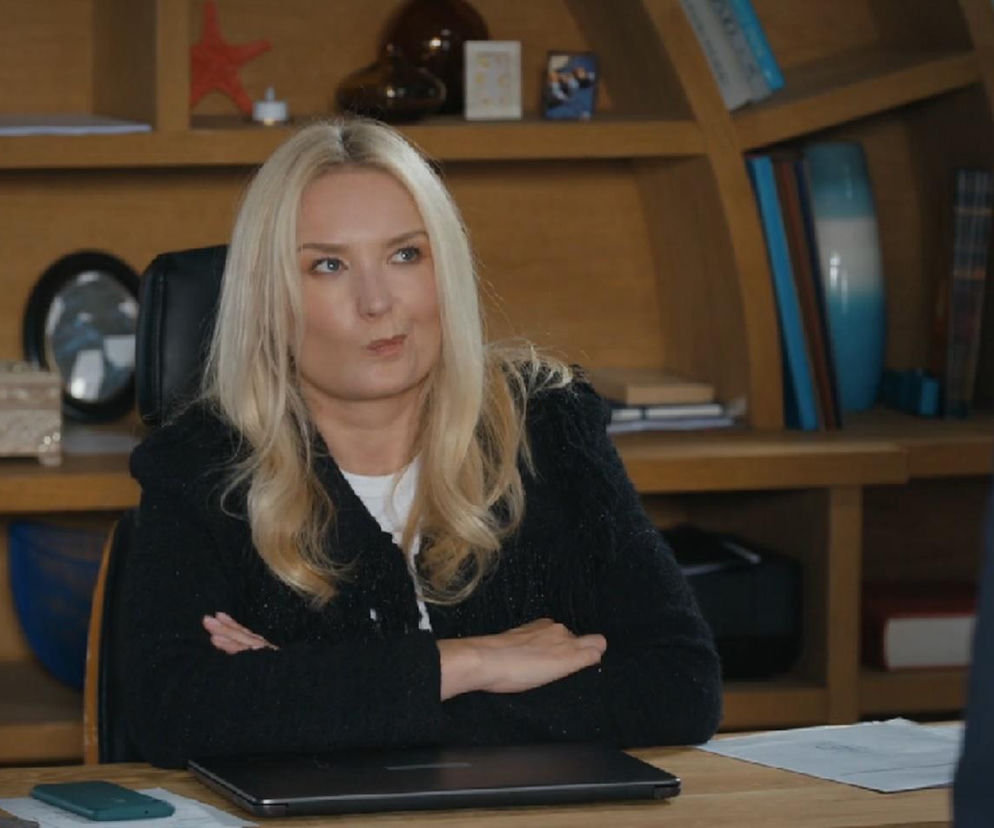 Marta Dąbrowska jako Karolina w serialu Barwy szczęścia siedzi przy biurku z założonymi rękami, wyrażając niezadowolenie. Na biurku widoczny jest laptop i dzwonek, a w tle regał z książkami i ozdobami. Więcej o wątku Karoliny przeczytasz na Super Seriale.