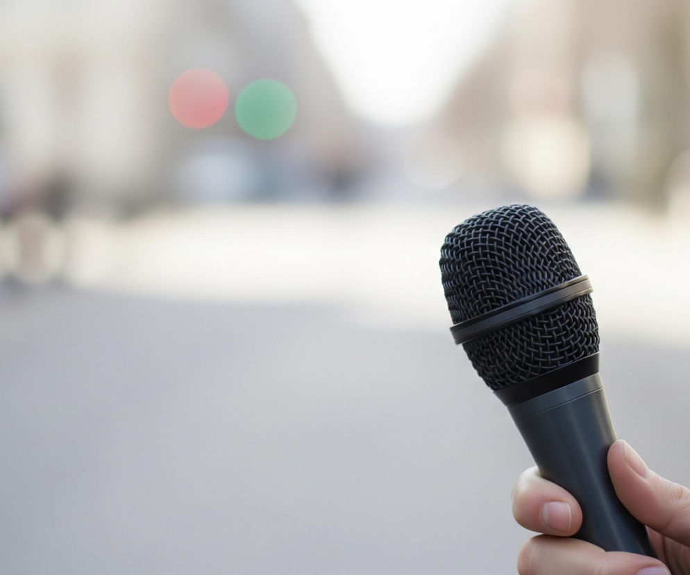 Prawa dłoń trzyma ciemnoszary mikrofon dynamiczny z czarną, siateczkową osłoną, skierowany w górę i lekko w lewo. Tło jest rozmyte, przedstawiające ulicę z rozmytymi budynkami i sylwetkami ludzi, w tym dwoma okrągłymi światłami w kolorze czerwonym i zielonym, sugerującymi sygnalizację świetlną. Całość utrzymana jest w jasnej, szarej kolorystyce z delikatnymi akcentami barwnymi.