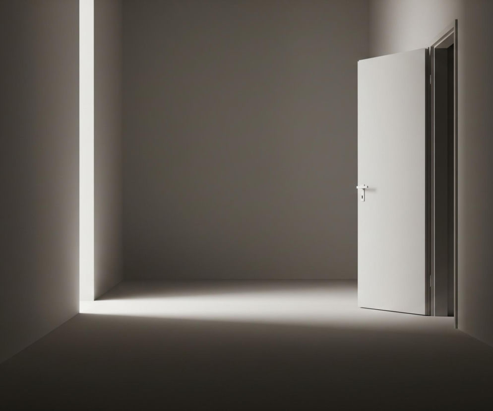 Pusta, szara przestrzeń, prawdopodobnie korytarz, rozjaśniona jest smugą jasnego światła wpadającego z lewej strony oraz przez lekko uchylone, jasne drzwi po prawej. Na podłodze widać wyraźny, świetlisty prostokąt, który biegnie od lewej ściany, rozjaśniając centralną część pomieszczenia. Uchylone drzwi ukazują ciemne wnętrze za nimi, a ich srebrna klamka wyraźnie odcina się na tle białej powierzchni.