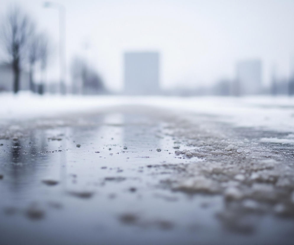 Zbliżenie na kałużę roztopionego śniegu i lodu na powierzchni drogi w zimowy dzień. Woda w kałuży odbija światło nieba, a na jej powierzchni unoszą się niewielkie grudki lodu i błota. Po obu stronach kałuży leży biały, nierówny śnieg. W oddali, niewyraźne, widać drzewa bez liści, słupy oraz zarysy budynków.