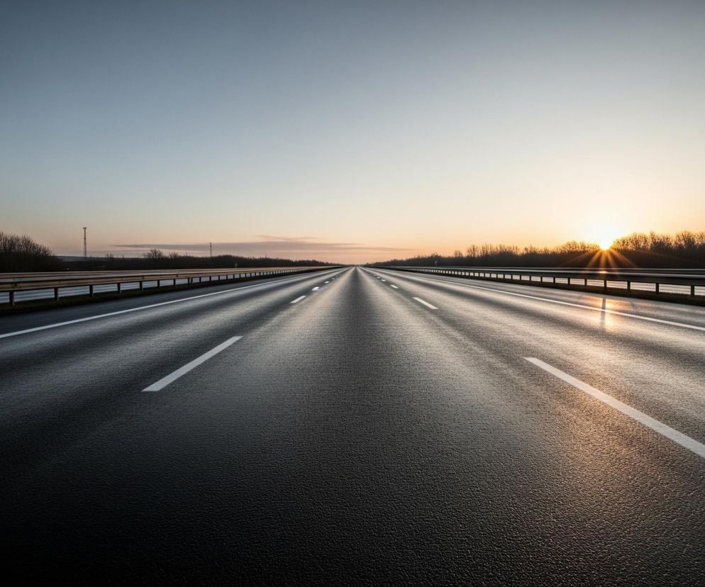 Czarna, pusta autostrada rozciąga się w głąb kadru, z dwoma pasami ruchu w każdym kierunku, oddzielonymi białymi liniami. Nawierzchnia drogi jest mokra i odbija światło w centralnej części. Horyzont przecina ciemny pas drzew, a za nim wschodzi słońce, malując niebo odcieniami pomarańczowego i niebieskiego. Po obu stronach autostrady widoczne są metalowe barierki ochronne.