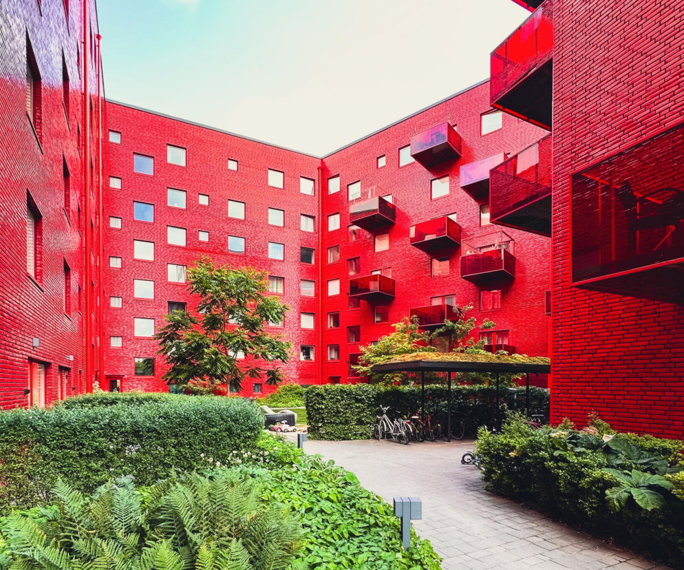 Czerwony budynek Muddus Kvarteret w Sztokholmie z intensywnie czerwonej, szkliwionej cegły, otaczający zielony dziedziniec z drzewami, krzewami i parkingiem rowerowym. Więcej o tej architekturze przeczytasz na Architektura Murator Plus.