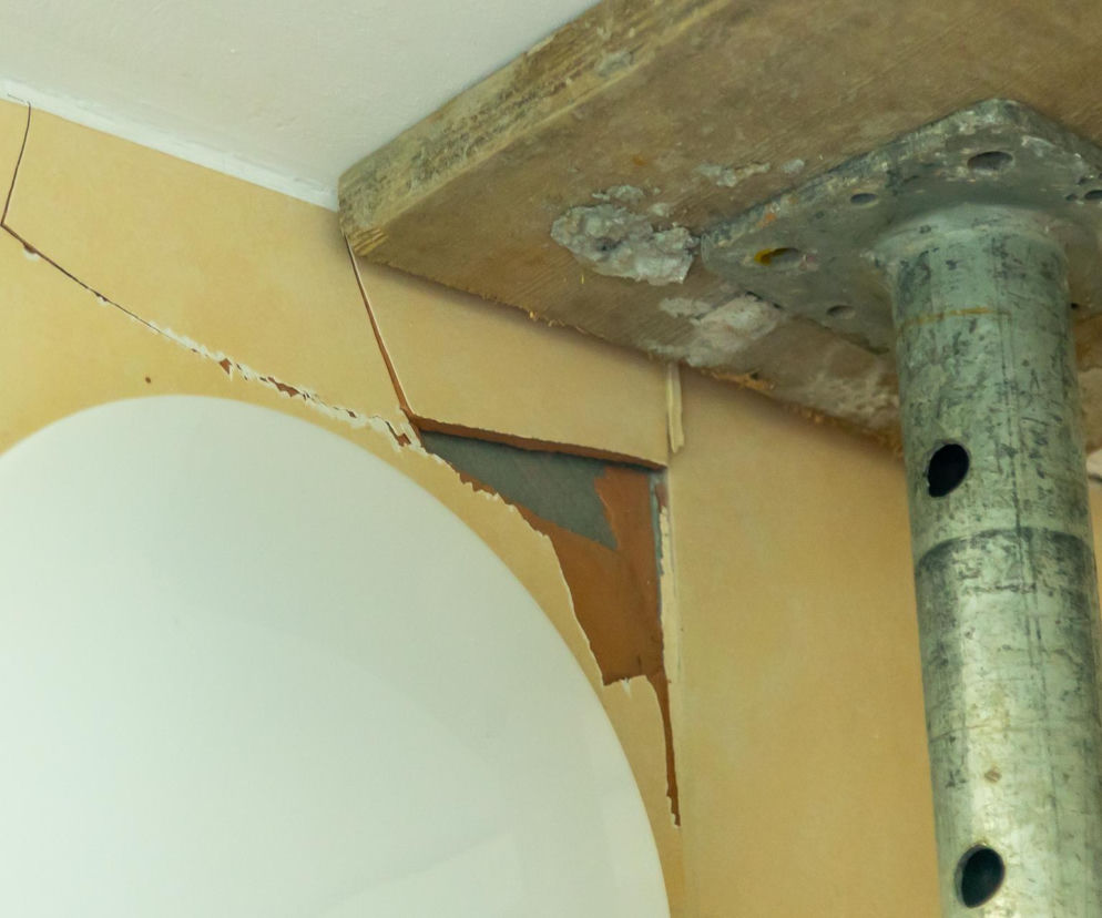 Zniszczona ściana z widocznymi pęknięciami i łuszczącą się farbą, wsparta metalowym stemplem budowlanym, co świadczy o problemach konstrukcyjnych bloku przy Hirszfelda 2 w Warszawie. Więcej o architekturze budynków możesz przeczytać na Architektura Murator Plus.