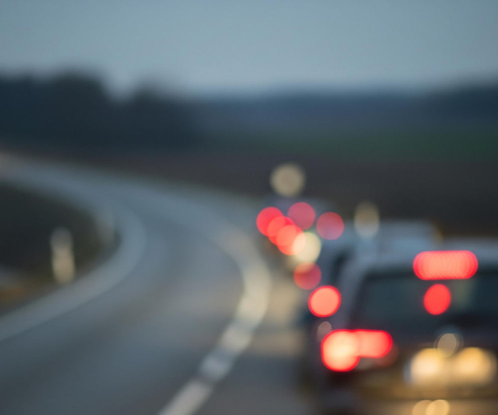 Droga biegnie w głąb obrazu, łagodnie skręcając w lewo, z rozmazanymi światłami samochodów tworzącymi efekt bokeh. Na pierwszym planie po prawej stronie widać tył samochodu z wyraźnymi czerwonymi światłami stopu i drogowymi, oraz słabszym, żółtawym światłem tablicy rejestracyjnej. Za nim, w rzędzie, znajdują się kolejne samochody, których tylne światła tworzą kaskadę czerwonych i pomarańczowych plam. Po lewej stronie, daleko w perspektywie, widoczne są dwa jasne, żółte punkty, prawdopodobnie światła nadjeżdżającego pojazdu, na tle niewyraźnego, ciemnego krajobrazu i zachmurzonego nieba.