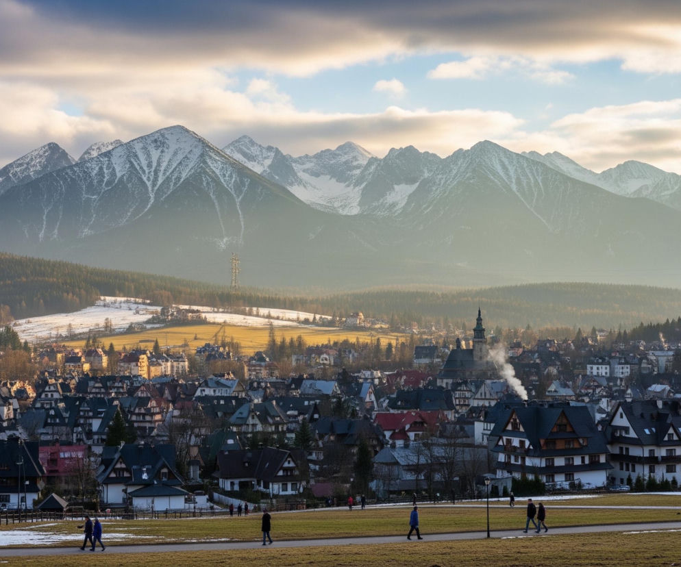 W oddali widoczne są majestatyczne, zaśnieżone pasma górskie o ostrych, skalistych szczytach, częściowo skryte w lekkiej mgiełce. Poniżej rozciąga się miejski krajobraz z licznymi domami o ciemnych, spadzistych dachach i jasnych elewacjach, wśród których wyróżnia się wieża kościoła oraz kominy dymiące białym dymem. Na pierwszym planie znajduje się szeroka, pokryta trawą przestrzeń z brązowymi ścieżkami, po których spacerują pojedyncze osoby i małe grupy. Niebo jest częściowo zachmurzone, z ciemniejszymi chmurami na górze i jaśniejszymi, rozświetlonymi przez słońce obłokami w centralnej części.