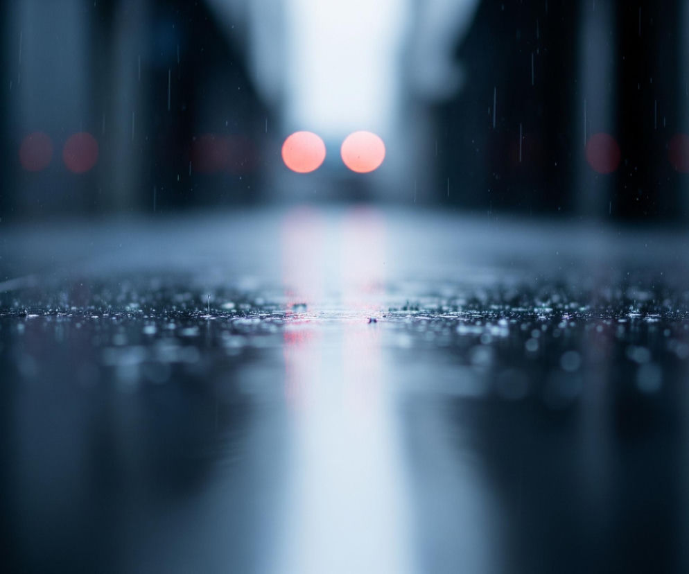 Ciemna, rozmyta droga pokryta jest drobnymi kroplami deszczu, które odbijają blade światło, tworząc efekt iskrzenia na powierzchni. W oddali, na końcu drogi, widoczne są dwa czerwone, rozmyte światła, prawdopodobnie tylne światła pojazdu, oraz jasna, biała przestrzeń. Całość utrzymana jest w chłodnej palecie barw, dominują ciemne odcienie szarości i błękitu, z elementami rozmytego światła tworzącego odbicia na mokrej nawierzchni.