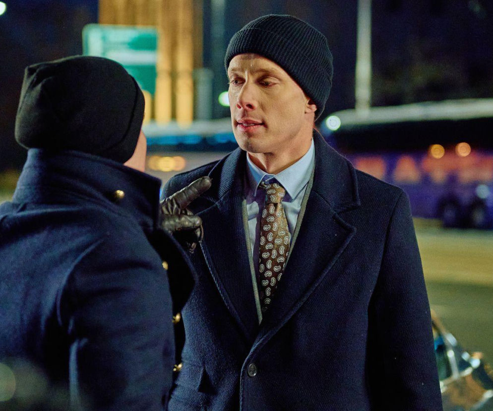 Aktor Darek Żbik w ciemnej czapce i płaszczu rozmawia z drugą osobą, wskazując palcem, podczas nocnej sceny z serialu Na Wspólnej. W tle widać rozmazane światła miasta i niebieski autobus. Dowiedz się więcej o serialu i dramatycznych wydarzeniach w Super Seriale.