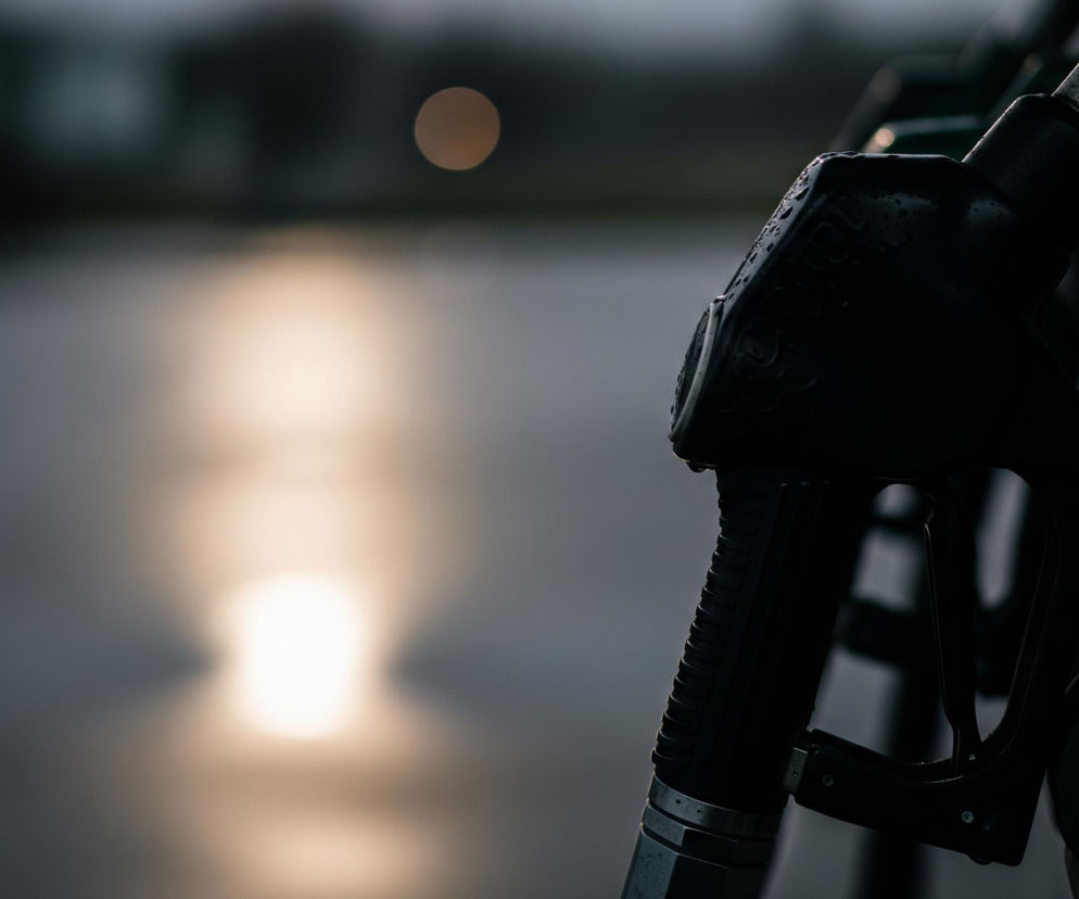 Ciemna dysza dystrybutora paliwa, pokryta kroplami wody, zajmuje prawą część obrazu, lekko odchylona w lewo. Jej powierzchnia jest czarna, z metalowymi elementami u podstawy oraz na lufie. W tle, lewej strony, widoczna jest rozmyta, jasna plama światła, odbijająca się od szarej, wodnistej powierzchni, a nieco wyżej po lewej stronie znajduje się mniejszy, okrągły punkt światła. Całość utrzymana jest w ciemnej, stonowanej kolorystyce, z dominującymi odcieniami szarości i czerni.