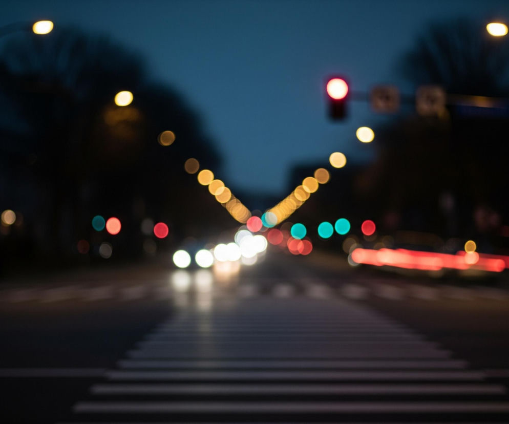 Na pierwszym planie widoczna jest ciemna ulica z wyraźnymi, białymi pasami przejścia dla pieszych. W tle rozciąga się miejski krajobraz nocą, z rozmytymi, kolorowymi światłami tworzącymi efekt bokeh. Dominują jasne, ciepłe światła uliczne w odcieniach żółci i pomarańczy, ułożone w rząd prowadzący w głąb obrazu. Po bokach widoczne są pojedyncze, czerwone światła sygnalizacji świetlnej oraz kilka zielonych, a także rozmyte smugi czerwonych świateł, przypominające tylne światła jadących samochodów, oraz jaśniejsze, białe plamy świateł z przodu pojazdów. Całość zanurzona jest w ciemnoniebieskim zmierzchowym niebie i czerni nocy.