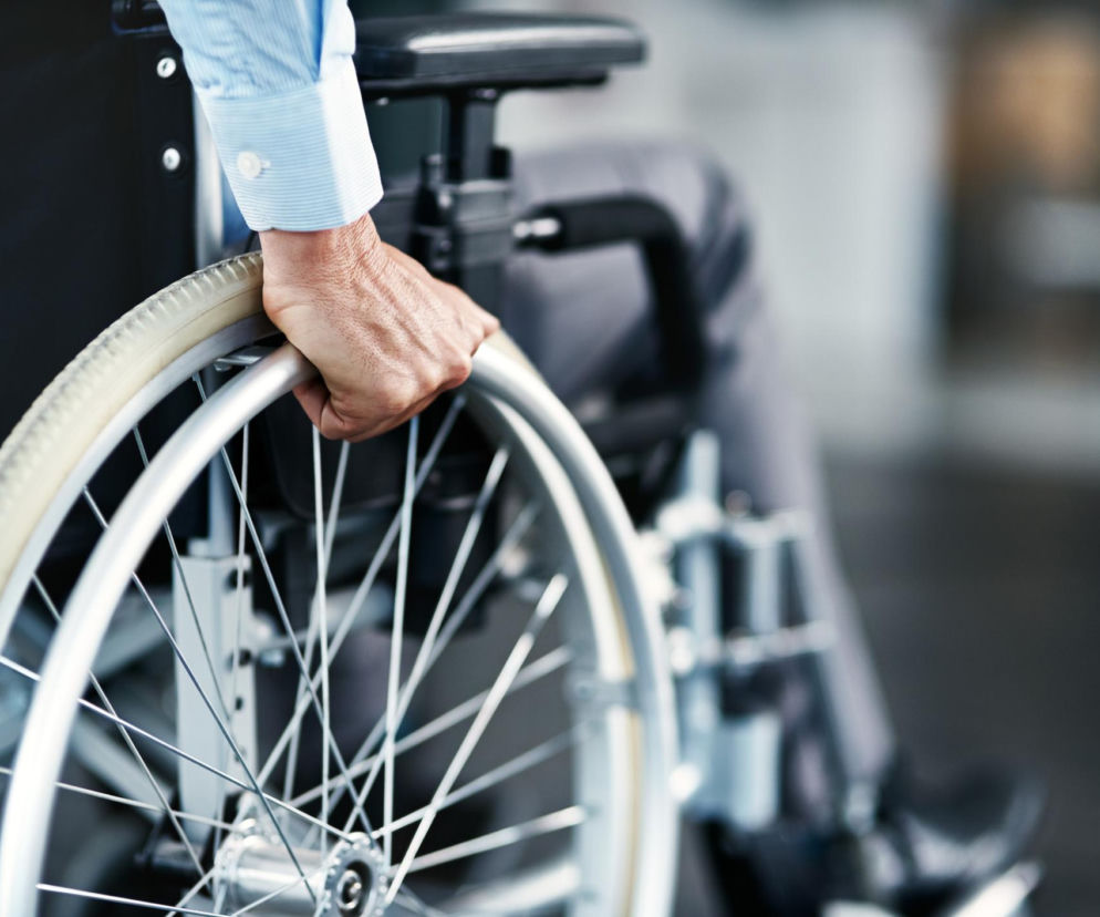 Ręka osoby w niebieskiej koszuli z mankietem, popychająca koło wózka inwalidzkiego. Obraz symbolizuje samodzielność i rehabilitację osób z niepełnosprawnością, o czym przeczytasz więcej na portalu Poradnik Zdrowie. Tło jest rozmyte.