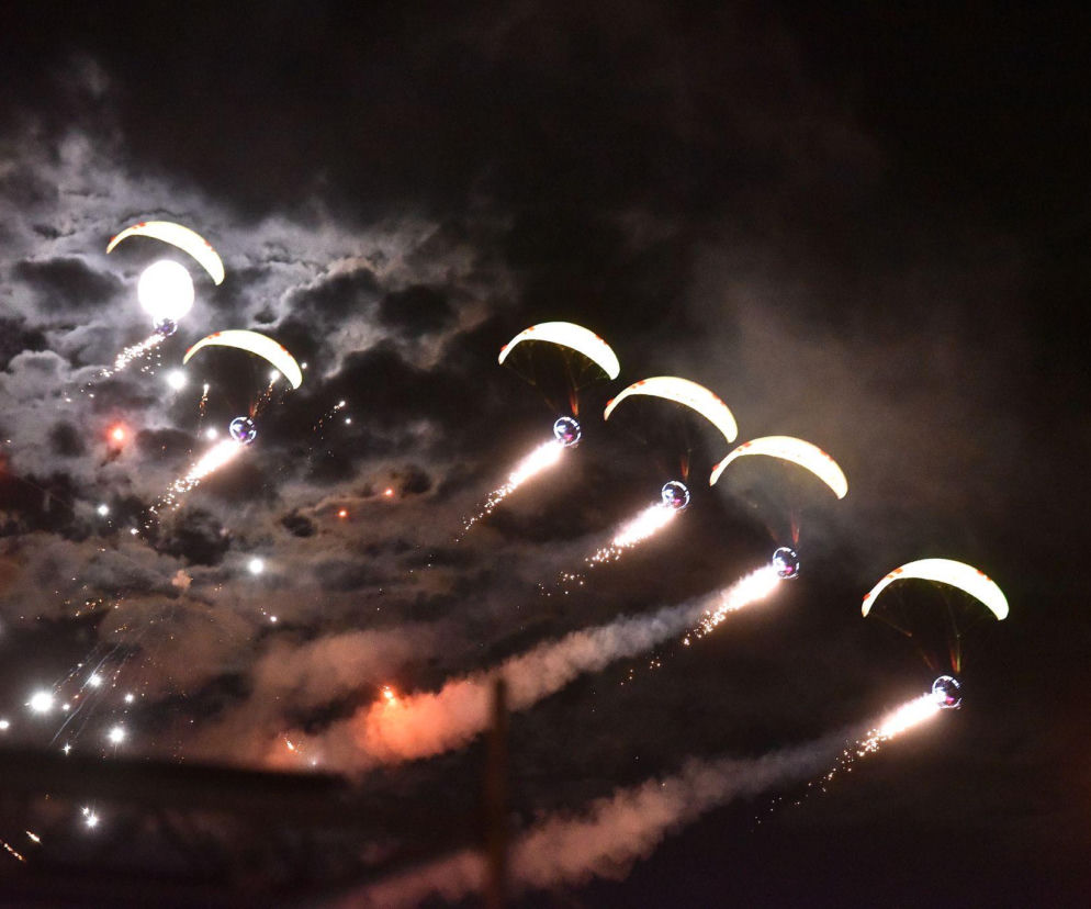 Sześciu paralotniarzy z oświetlonymi czaszami i pirotechniką leci na nocnym niebie. O Airshow przeczytasz na Eska Toruń.