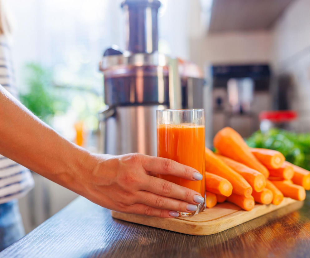 Kobieta w kuchni przygotowująca świeży sok z marchwi, obok sokowirówka i świeże marchewki na desce. Artykuł na Poradnik Zdrowie wyjaśnia, czy marchew naprawdę poprawia wzrok.