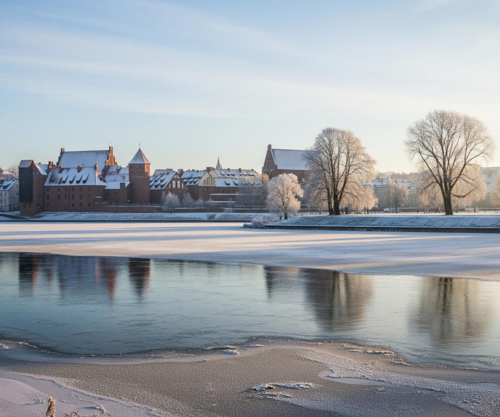 Zamarznięta rzeka w centrum kadru odbija niebieskie niebo i odległe, ośnieżone budynki, z ciemniejszą, otwartą wodą w środku. Na pierwszym planie, po lewej stronie, widać gałęzie drzew pokryte szronem. Horyzont tworzą czerwone, ceglane budowle z białymi, zaśnieżonymi dachami, oraz jasne drzewa również pokryte szronem, na tle bezchmurnego, jasnoniebieskiego nieba.