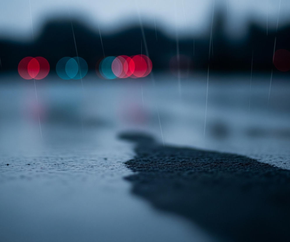 Na pierwszym planie widoczna jest mokra powierzchnia, prawdopodobnie asfalt, z wyraźnymi kroplami deszczu lub spływającą wodą, tworzącą drobne zmarszczki na tafli. Prawa strona obrazu, na pierwszym planie, ukazuje bardziej szorstką, suchą teksturę podłoża, graniczącą z mokrą powierzchnią, co tworzy wyraźny kontrast. W tle, rozmyte światła samochodowe w kolorach czerwonym i niebieskim, odbijają się w kałużach, tworząc świetliste smugi i kręgi. Całość utrzymana jest w chłodnych, ciemnych odcieniach szarości i błękitu, z pionowymi, jasnymi liniami symulującymi padający deszcz.