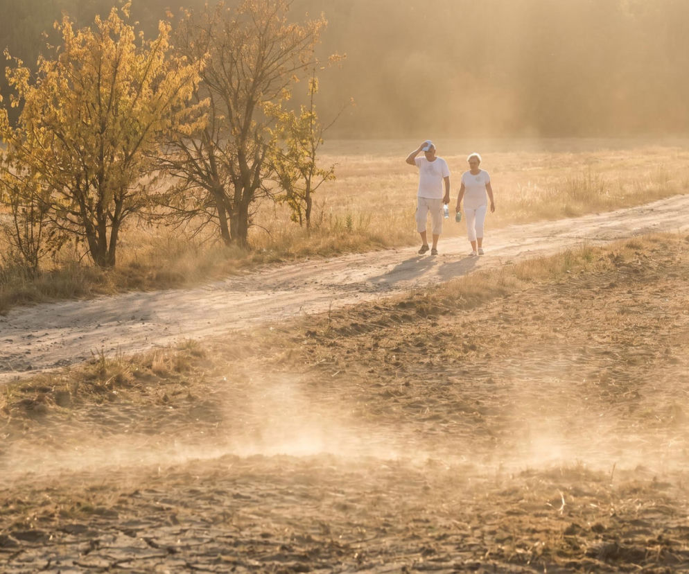 Para seniorów w białych ubraniach idzie piaszczystą drogą w upalny dzień, unoszący się kurz symbolizuje wysokie temperatury. O zagrożeniach związanych z rekordowymi upałami przeczytasz więcej na naszym portalu.