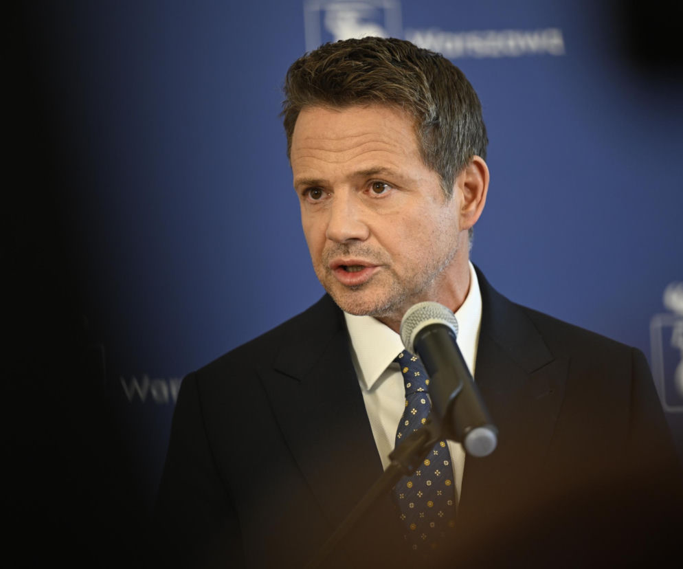 Rafał Trzaskowski przemawiający do mikrofonu na tle granatowej ściany z logo miasta, w trakcie dyskusji o aferze Szpitala Południowego i jego możliwej dymisji. Więcej o kryzysie politycznym przeczytasz na naszym portalu.