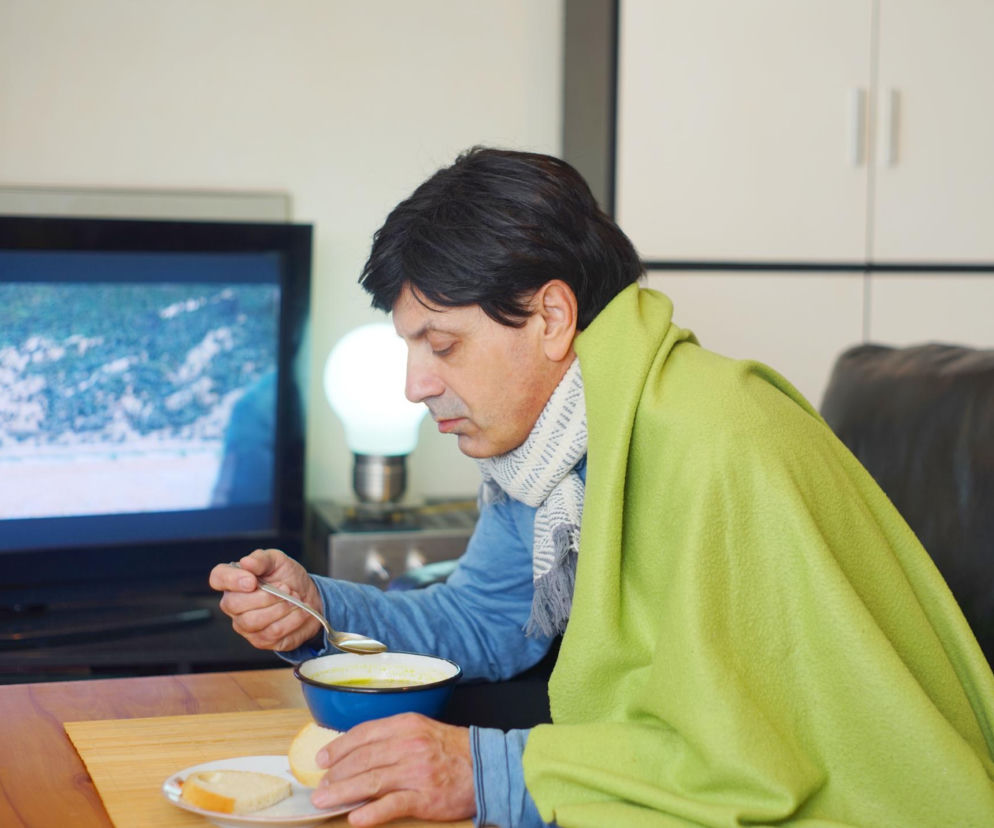 Mężczyzna w średnim wieku, okryty jasnozielonym kocem i szarym szalikiem, je zupę z niebieskiej miski. Obok leżą kromki chleba. Scena w domu, symbolizująca dreszcze i uczucie chłodu po jedzeniu, o czym możesz przeczytać na portalu Poradnik Zdrowie.