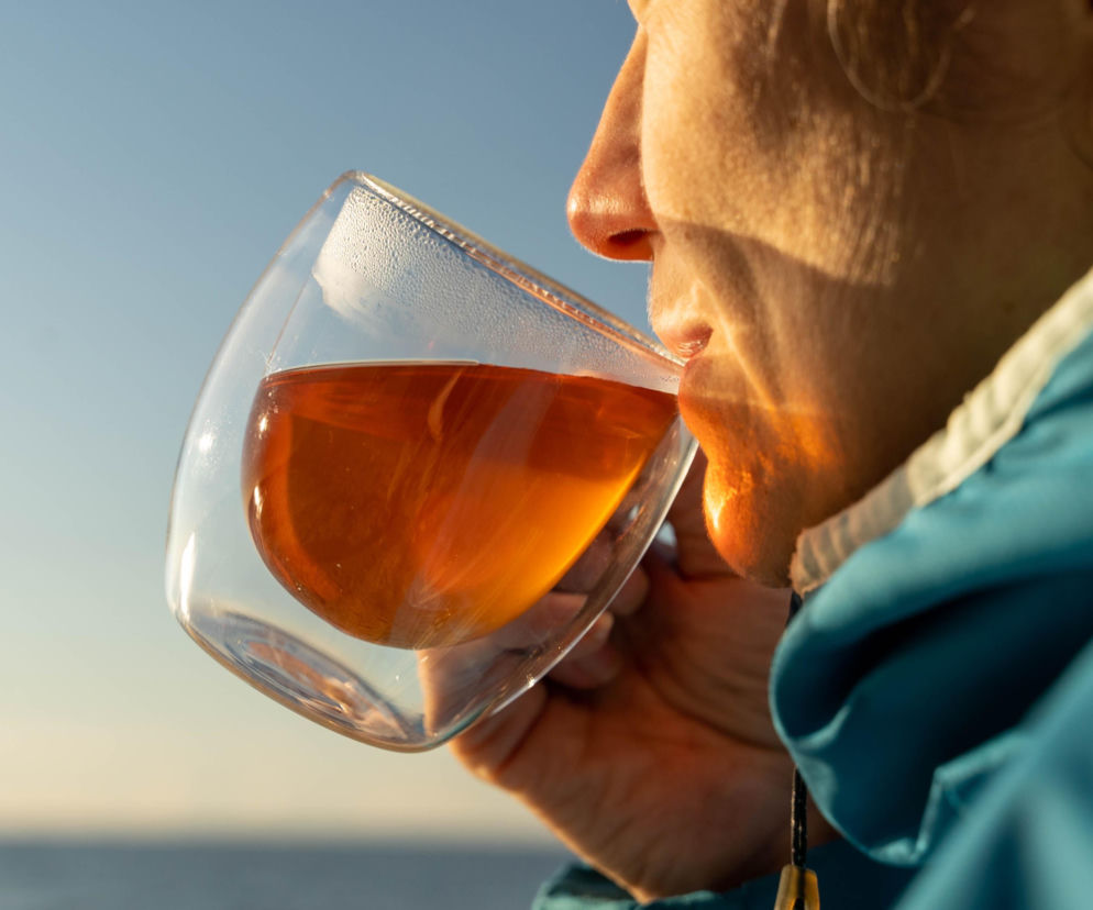 Kobieta pijąca herbatę rooibos z przezroczystego kubka z podwójnymi ściankami na tle błękitnego nieba i morza. Jej twarz jest oświetlona ciepłym, bocznym światłem. Dowiedz się więcej o korzyściach płynących z picia herbaty rooibos na naszym portalu.