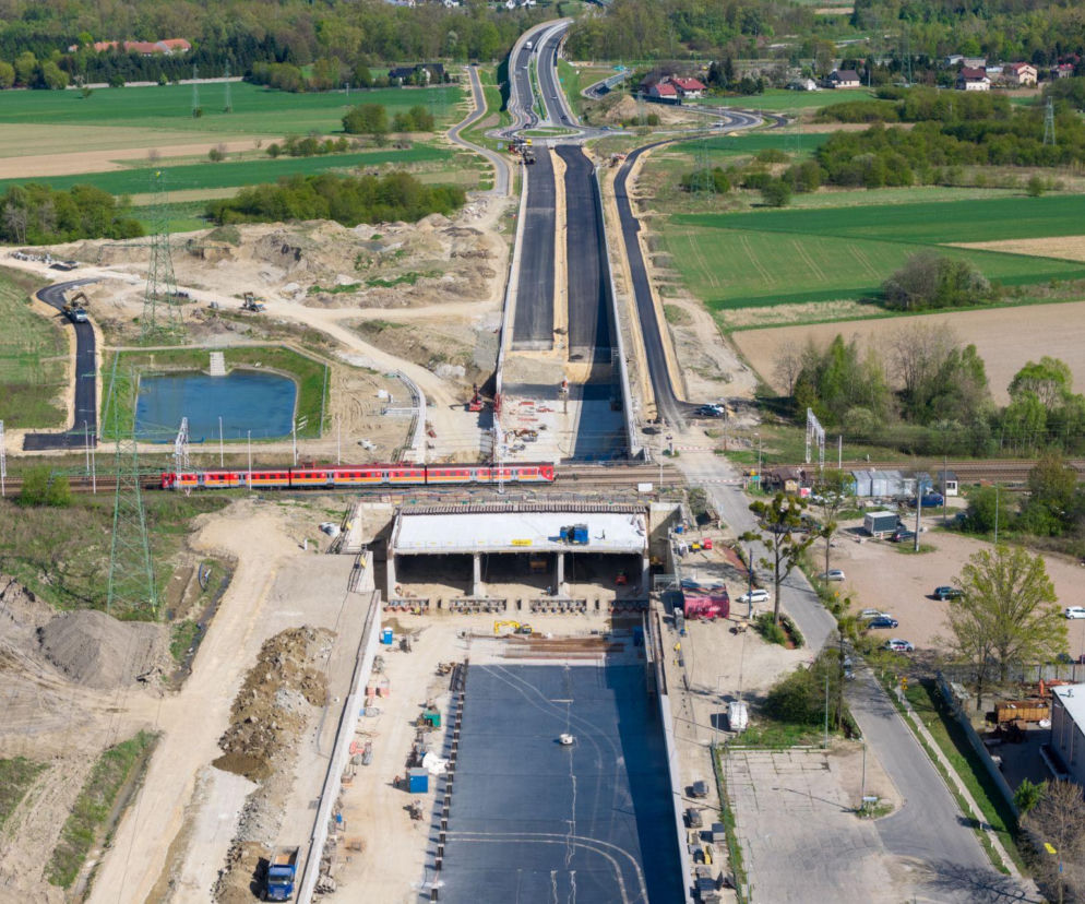 Widok z lotu ptaka na budowę obwodnicy Oświęcimia, z widocznym betonowym prostopadłościanem wciskanym pod tory kolejowe, nad którymi przejeżdża czerwony pociąg. Nowoczesne rozwiązania inżynieryjne stosowane na tej budowie są opisane na portalu Architektura Murator Plus.