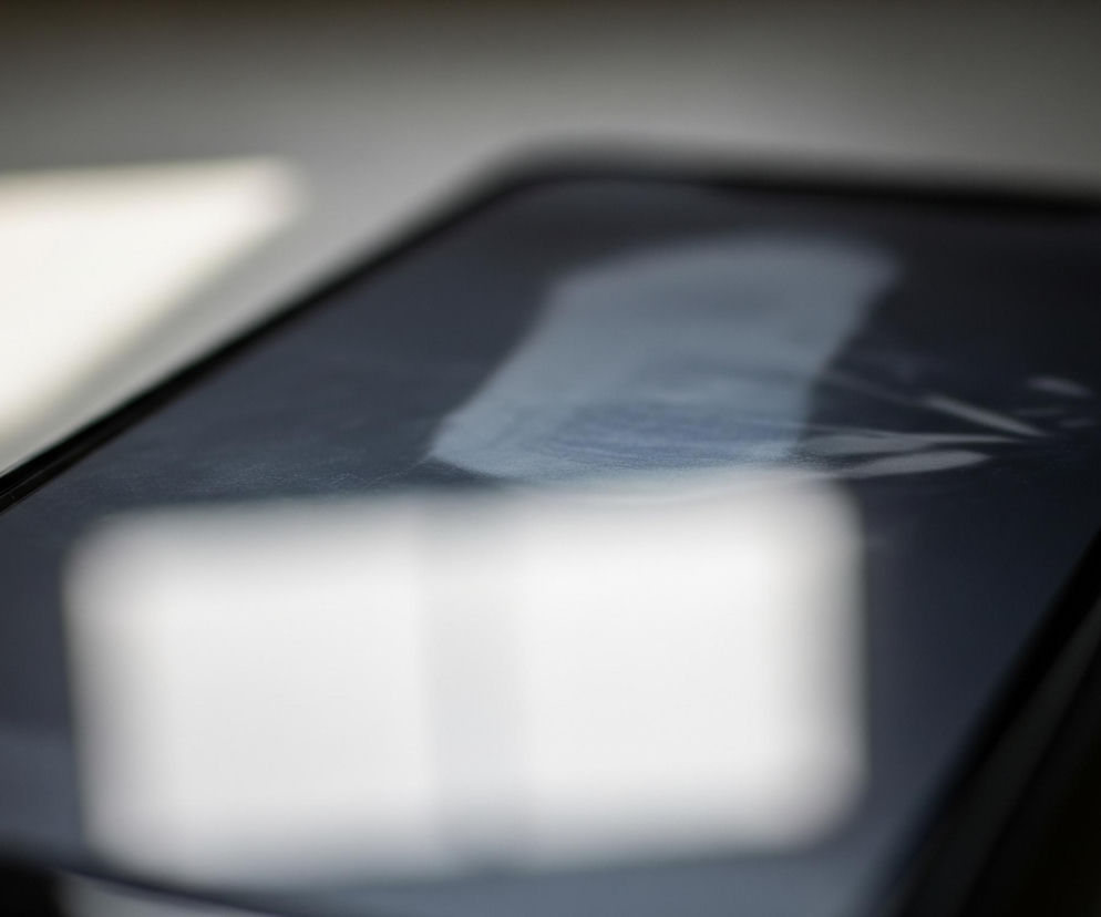 Na pierwszym planie leży smartfon, ujęty pod kątem, z lewej strony nieco bliżej do obiektywu. Jego czarny ekran odbija rozmyte, jasne światło w kształcie prostokąta, z widoczną w górnej części smugą światła, przypominającą odbicie okna lub lampy. Obudowa telefonu jest czarna, a po prawej stronie widać lekko wystający, ciemny przycisk. Tło za smartfonem jest jasnoszare, z dwoma rozmazanymi, białymi prostokątami, które są odbiciami źródła światła, podobnymi do tych na ekranie.