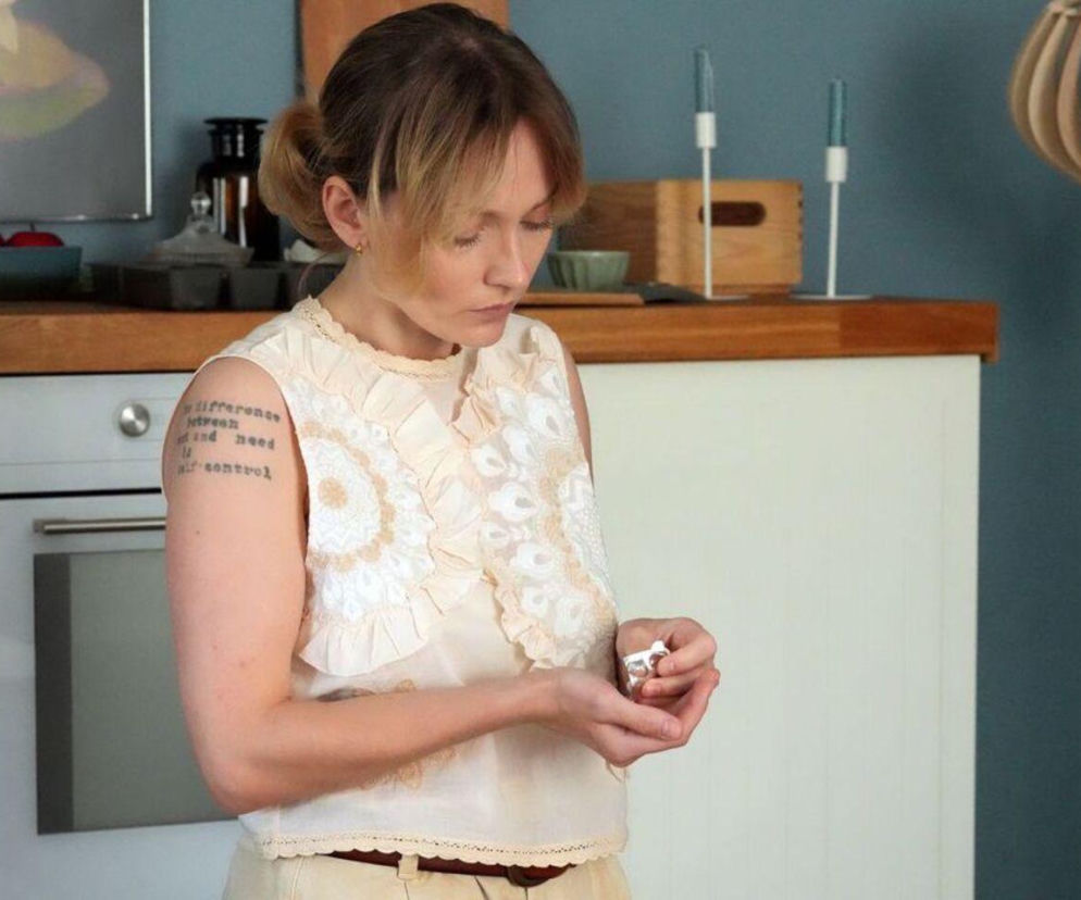Renata (Anna Mrozowska) w jasnej bluzce z falbanami, stojąca w kuchni i trzymająca w dłoniach blister z tabletkami. Aktorka z serialu Barwy szczęścia ma na ramieniu tatuaż. Więcej o stanie zdrowia Renaty przeczytasz na portalu Super Seriale.
