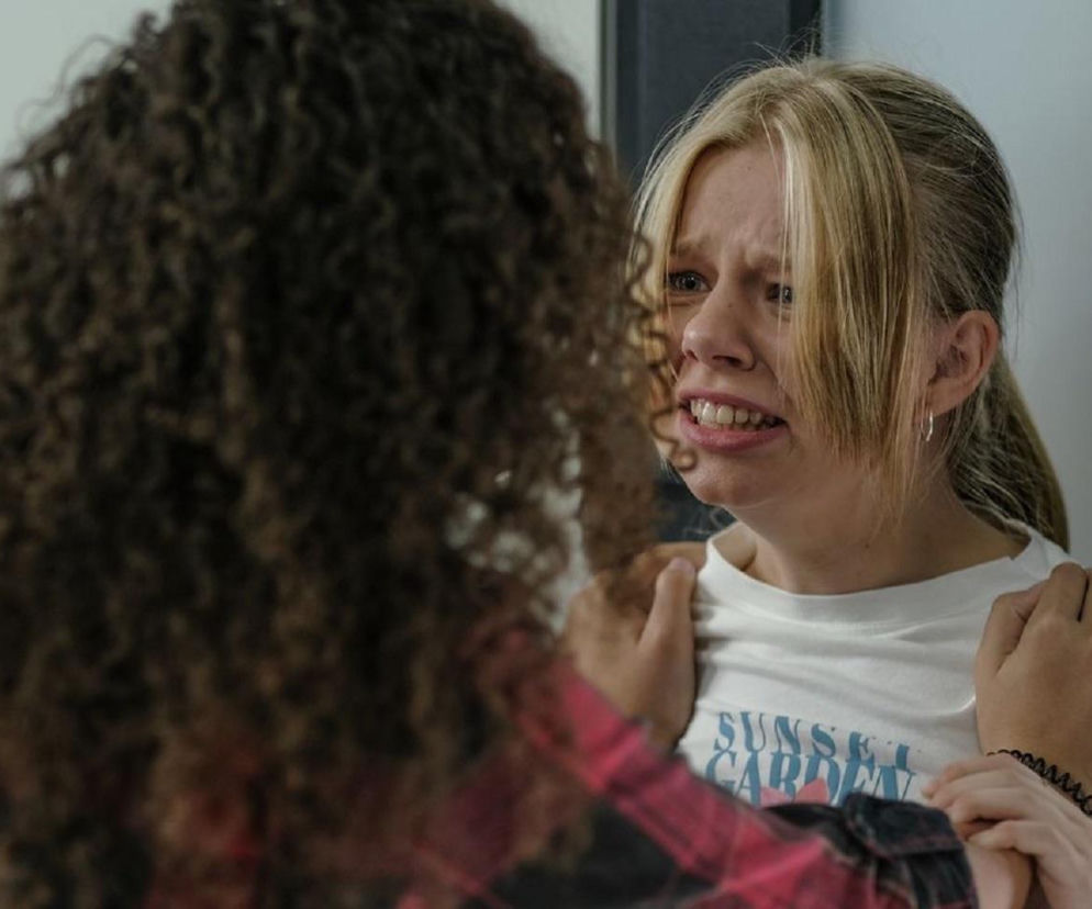 Przerażona Lenka ze łzami w oczach, z blond włosami spiętymi w kucyk, w białej koszulce z niebieskim napisem, stoi z szeroko otwartymi ustami, wyrażając swój szok i strach. W objęciach trzyma ją tajemnicza postać z kręconymi, brązowymi włosami, której plecy widać. Więcej informacji o serialu M jak miłość znajdziesz na Super Seriale.