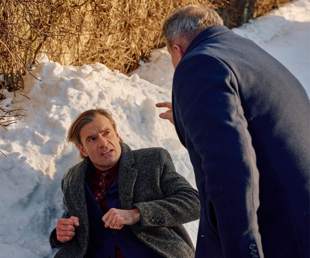 Mariusz Czerski, ubrany w płaszcz i marynarkę, siedzi na śniegu z wyrazem złości i desperacji, podczas gdy drugi mężczyzna, stojący tyłem, wymachuje do niego ręką. Scena bójki na cmentarzu z serialu Na Wspólnej w nowym odcinku, dostępna na stronie Super Seriale.