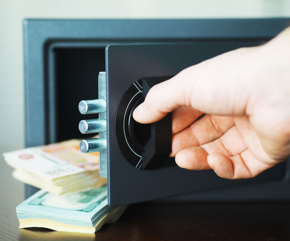 Otwierana dłonią czarna kasetka sejfu, z której wystają pliki banknotów, symbolizuje zabezpieczenie gotówki na wypadek kryzysu, o którym przeczytasz na Super Biznes. Banknoty w środku sejfu sugerują przechowywanie pieniędzy w domu jako formę zabezpieczenia finansowego.