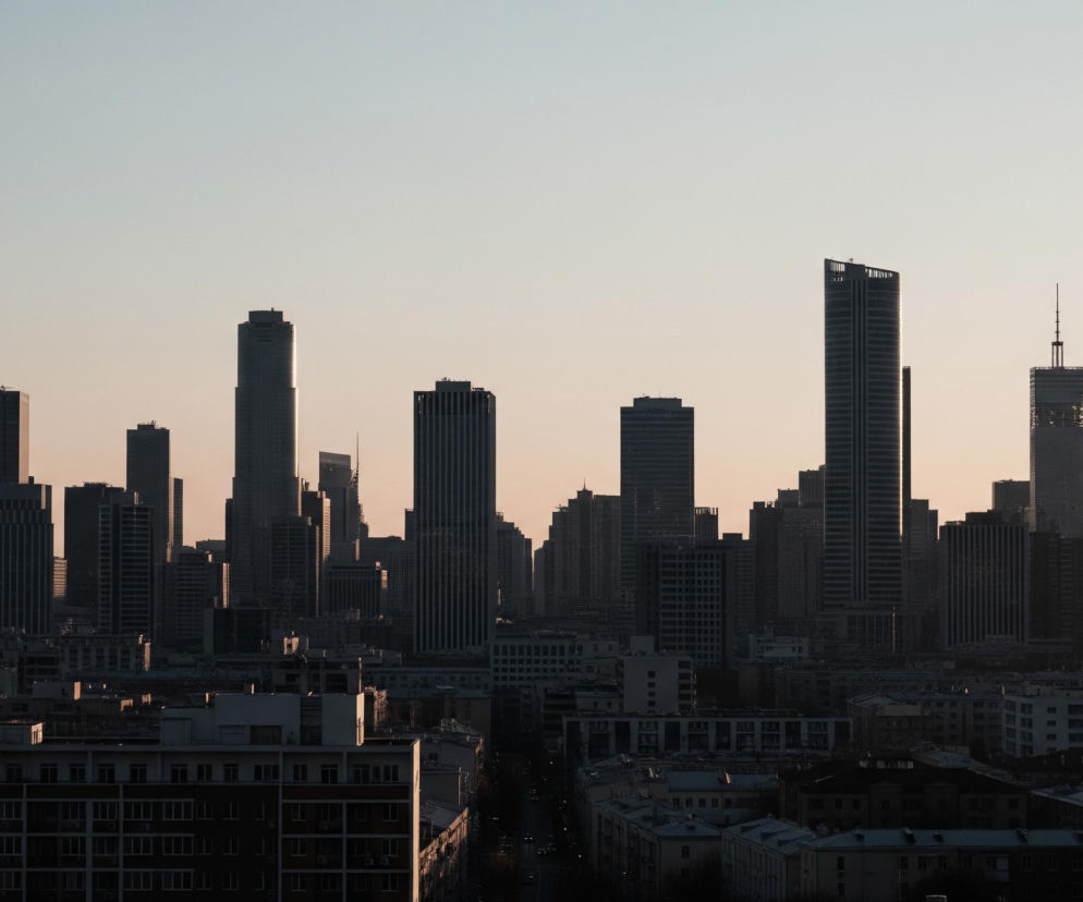 Panoramę miasta wypełnia horyzont wysokich, ciemnych budynków, z których najwyższe dominują w centrum kadru. Sylwetki drapaczy chmur są w większości zacienione, odcinając się ostro od jasnego, gradientowego nieba, które przechodzi od błękitu na górze do delikatnego pomarańczu przy horyzoncie. Na pierwszym planie widoczne są niższe budynki mieszkalne z białymi ścianami i ciemnymi dachami, częściowo oświetlone złotym światłem. Wśród nich biegnie ulica z rzędem słabo widocznych samochodów.