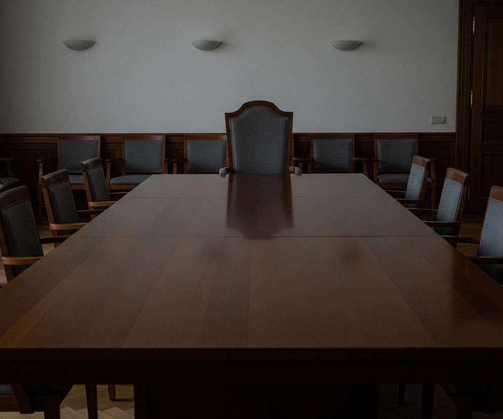 W centrum obrazu znajduje się długi, prostokątny stół konferencyjny wykonany z ciemnego, brązowego drewna o gładkim, błyszczącym blacie, który odbija światło z górnej lewej strony. Stół otoczony jest licznymi krzesłami z drewnianymi ramami i szarymi, tapicerowanymi siedziskami oraz oparciami, równomiernie rozmieszczonymi wzdłuż jego boków, a jedno większe, bardziej zdobione krzesło stoi na szczycie stołu. Pomieszczenie ma białe ściany, a dolna ich część wyłożona jest drewnianą boazerią; na lewej ścianie widoczne są jasne, pionowe zasłony oraz ciemniejsze, brązowe zasłony. Na ścianie nad stołem zawieszone są trzy owalne, szare kinkiety, a po prawej stronie widać ciemne, podwójne drzwi z mosiężną klamką.