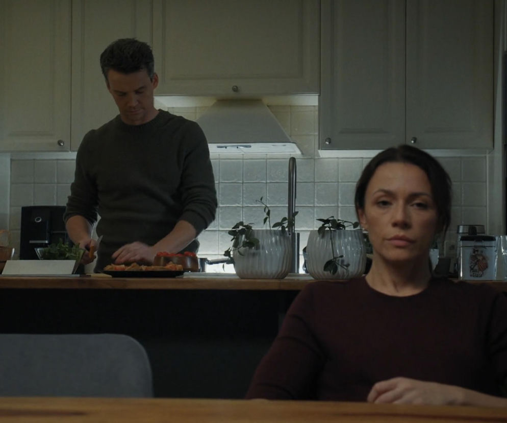 Aktorzy Krzysztof Kwiatkowski jako Jakub i Magdalena Turczeniewicz jako Martyna z serialu M jak miłość w scenie kuchennej. Jakub przygotowuje jedzenie, a Martyna siedzi przy stole, patrząc prosto. Więcej o ich relacji dowiesz się na Super Seriale.