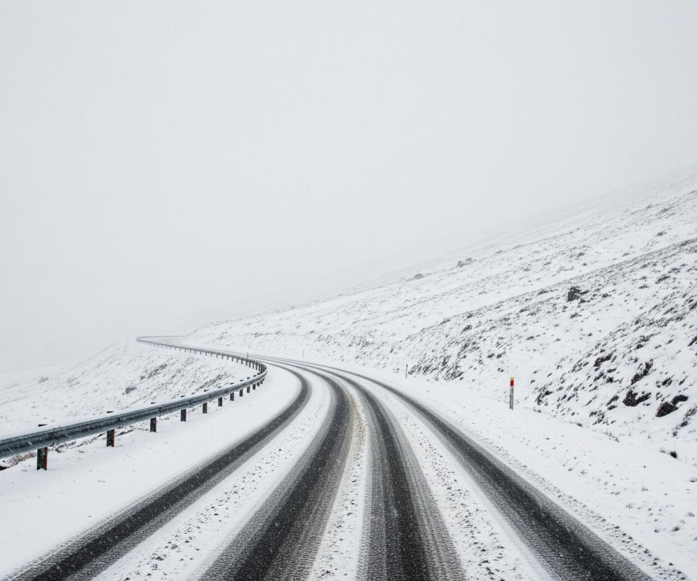 Droga pokryta śniegiem, z wyraźnymi czarnymi śladami kół, biegnie zakrętami w głąb obrazu, znikając w odległej, zamglonej bieli. Wzdłuż lewej krawędzi jezdni ciągnie się metalowa barierka ochronna osadzona na brązowych słupkach, również częściowo pokryta śniegiem. Prawa strona obrazu przedstawia zaśnieżone zbocze z widocznymi ciemnymi plamami roślinności lub skał, a przy krawędzi drogi stoi biało-czerwony słupek.
