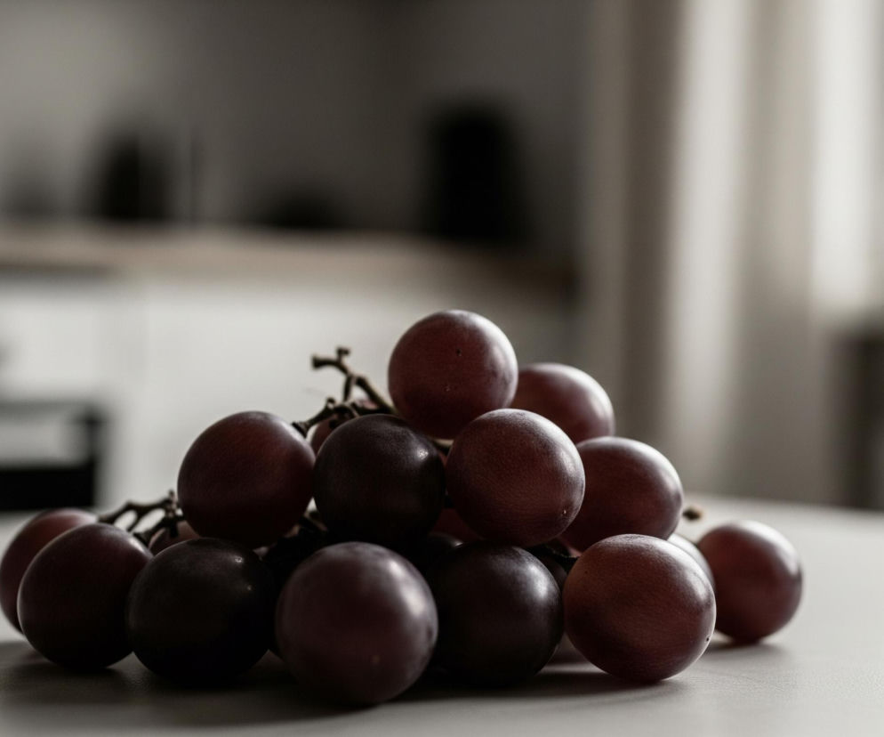 Grona czerwonych winogron leżą na jasnej, płaskiej powierzchni, zajmując centralną część kadru. Owoce mają ciemnoczerwony, niemal fioletowy odcień, a niektóre są ciemniejsze od innych, tworząc gradient kolorystyczny. Winogrona są zebrane w kiść, z widocznymi cienkimi, ciemnymi łodyżkami łączącymi poszczególne jagody. Tło jest rozmyte, utrzymane w odcieniach szarości i bieli, z widocznymi niewyraźnymi konturami mebli lub ścian, sugerującymi wnętrze pomieszczenia. Światło pada z prawej strony, oświetlając jaśniejsze partie winogron i tworząc delikatne cienie na powierzchni.