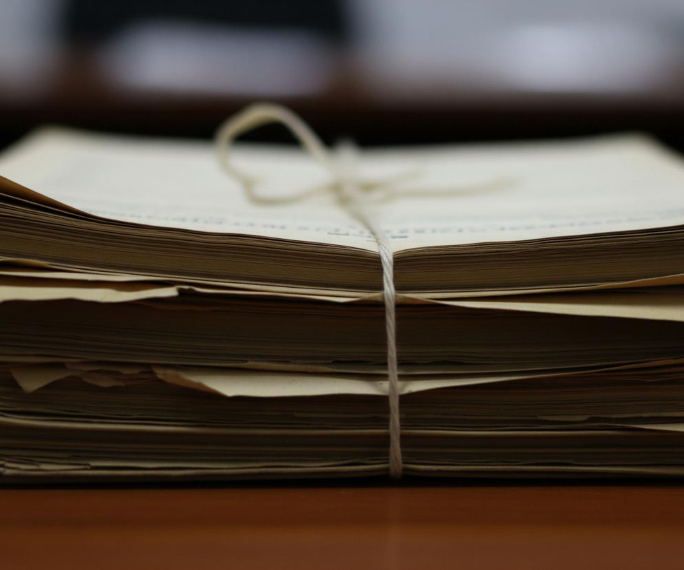 Na środku obrazu leży stos jasnobrązowych kartek papieru, ciasno związanych kremowym sznurkiem. Kartki są nieco pomarszczone i mają nierówne krawędzie, co sugeruje ich wiek lub częste użycie. Na górze stosu widać zawiązany sznurek tworzący pętelkę. Stos kartek spoczywa na gładkiej, brązowej powierzchni stołu lub biurka. Tło jest rozmyte, ciemniejsze, z jaśniejszymi fragmentami, co skupia uwagę na głównym obiekcie.