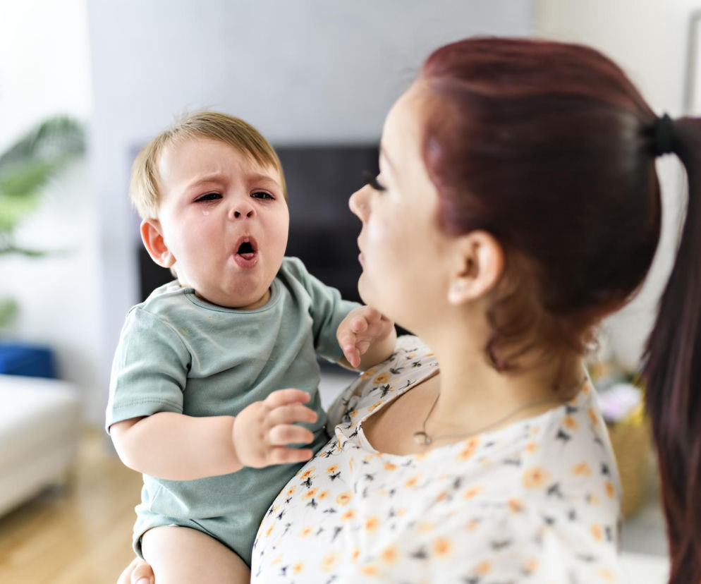 Kobieta trzyma płaczące niemowlę z otwartą buzią i łzami. O niepokojącym kaszlu u dzieci przeczytasz na MJM.