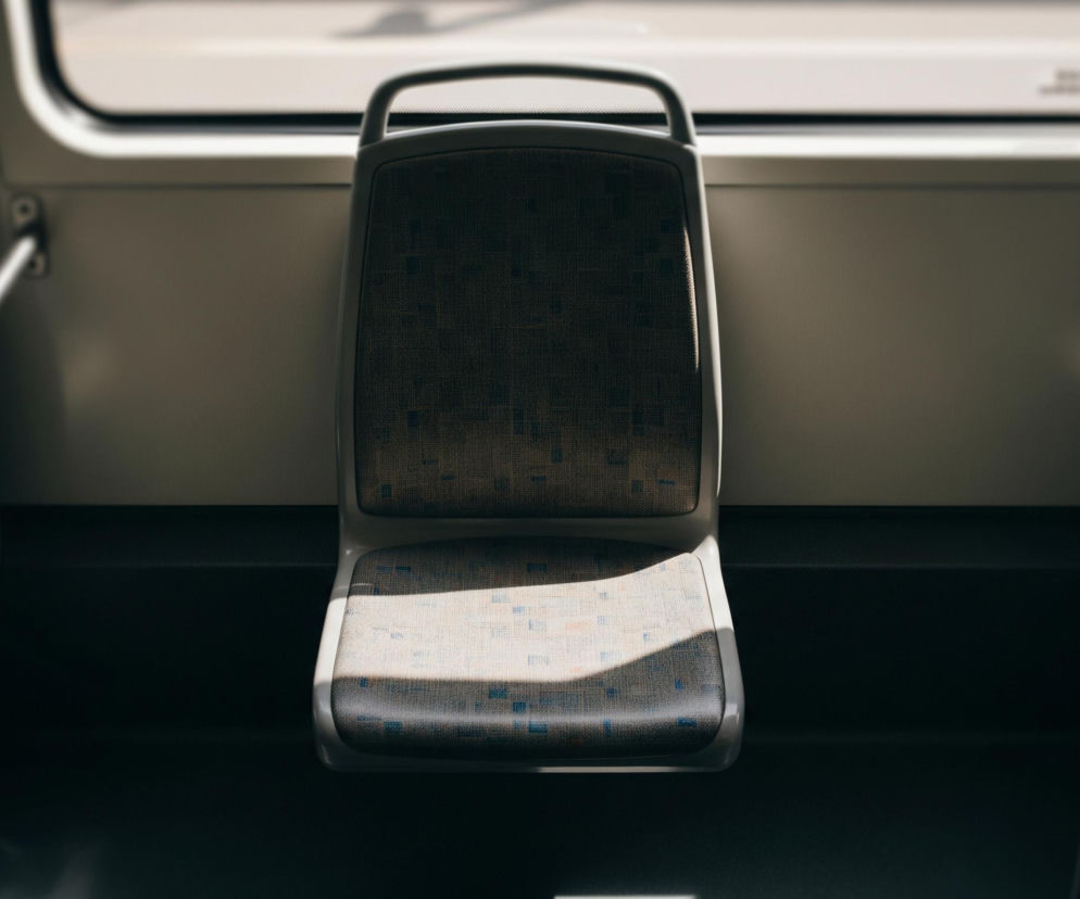 Centralnie na pierwszym planie widoczne jest puste siedzenie w pojeździe komunikacji miejskiej, prawdopodobnie tramwaju lub autobusie. Siedzenie ma szarą tapicerkę z drobnym, wzorem przypominającym piksele w odcieniach beżu i błękitu, z zaokrąglonymi krawędziami z jasnoszarego tworzywa. Pas światła słonecznego pada ukośnie na siedzisko, rozjaśniając jego prawą stronę i tworząc wyraźny cień na podłodze oraz częściowo na lewej stronie oparcia. W tle znajduje się okno, przez które wpada światło, a także fragmenty innych siedzeń i pionowych metalowych rur, służących jako uchwyty dla pasażerów, po obu stronach kadru.