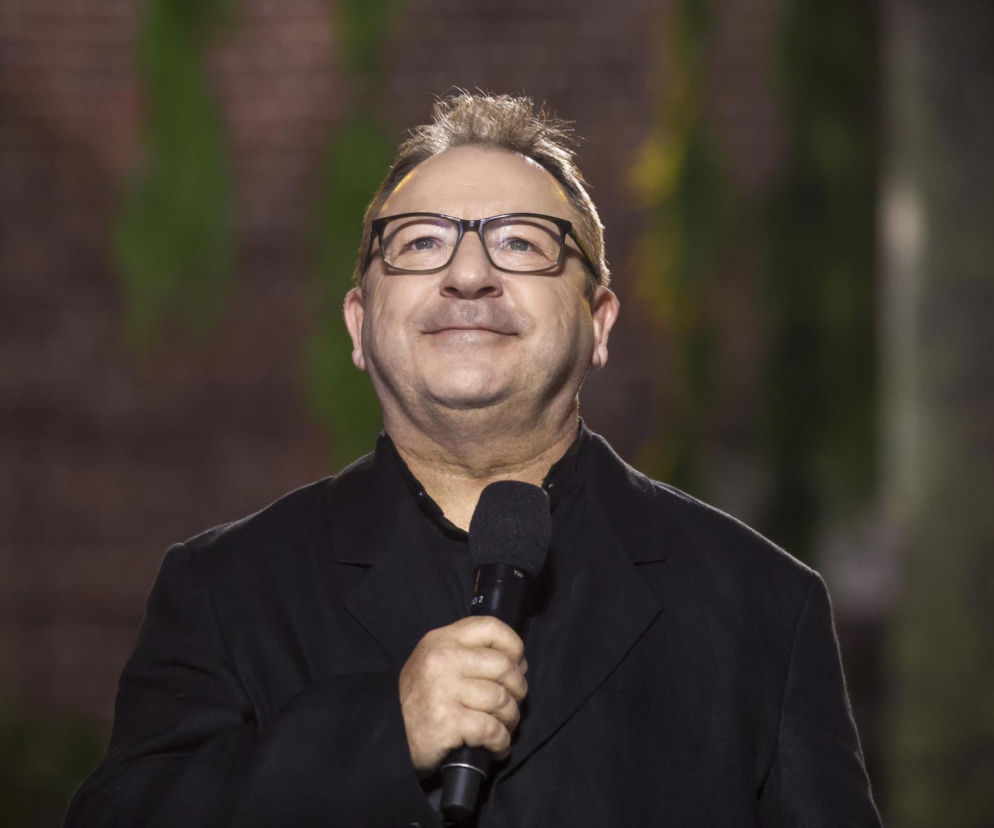 Aktor Zbigniew Zamachowski w okularach i czarnej marynarce trzyma mikrofon. O jego metamorfozie przeczytasz na SE.