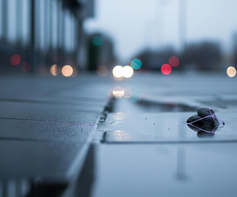 Na pierwszym planie, w prawym dolnym rogu, leży grupa ciemnoszarych kamieni związanych fioletową wstążką, częściowo zanurzonych w kałuży wody. Płytka, rozlana woda, z widocznymi refleksami światła, rozciąga się przez cały dół kadru i stanowi odbicie otoczenia. W tle rozmazane, kolorowe plamy światła – czerwone, żółte i turkusowe – tworzą efekt bokeh, sugerując ruch uliczny i światła miasta, rozmyty budynek znajduje się po lewej stronie.