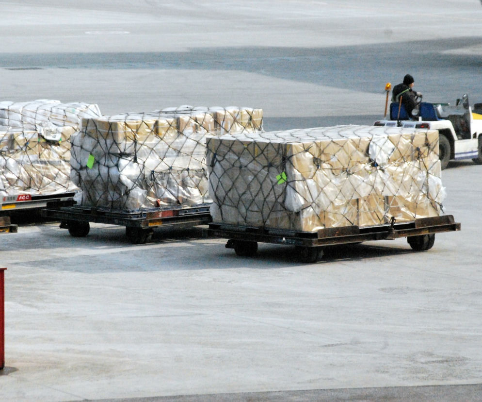 Ładunki cargo na wózkach bagażowych transportowane na płycie lotniska przez obsługę, symbolizujące globalną logistykę i przesyłki kurierskie. Artykuł na Super Biznes informuje o wejściu chińskiego giganta SF Express na polski rynek.