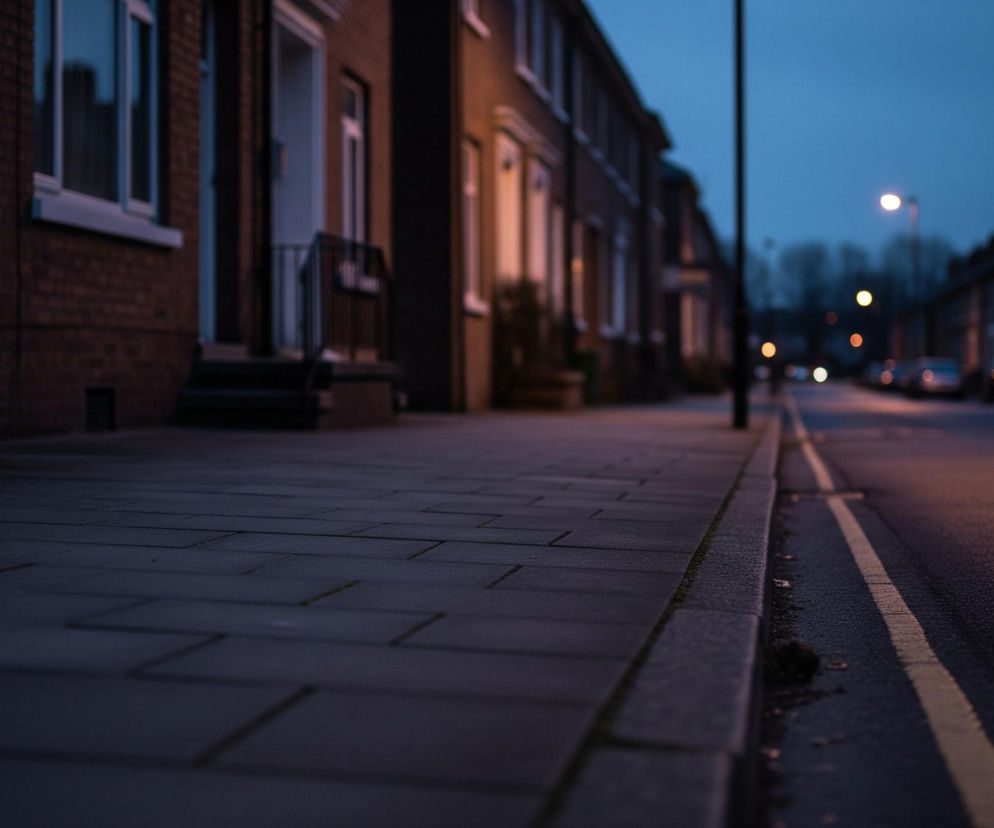 Ciemny chodnik z betonowych płyt biegnie wzdłuż lewej strony kadru, z delikatnie oświetlonymi fasadami ceglanych budynków po lewej stronie. Na prawo od chodnika znajduje się asfaltowa ulica z żółtą linią ciągłą, w oddali widoczne są światła uliczne i reflektory samochodów rozmywające się w wieczornej mgle. Niebo jest ciemnobłękitne, a ogólna atmosfera zdjęcia jest chłodna i spokojna.