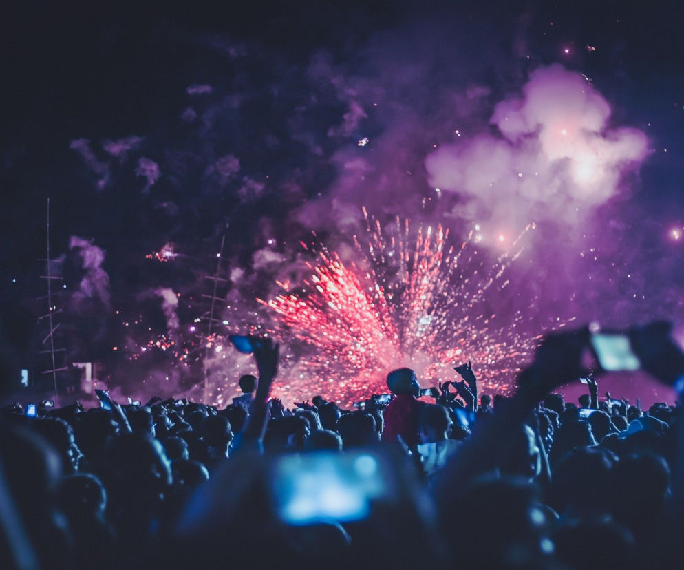 Tłum ludzi oglądający widowisko fajerwerków na ciemnym nocnym niebie. Rozbłyski czerwonych i fioletowych iskier rozświetlają chmury dymu, tworząc spektakularny widok. Wielu uczestników trzyma smartfony, rejestrując to wydarzenie. O skutkach nieostrożnej zabawy fajerwerkami przeczytasz na Super Biznes.