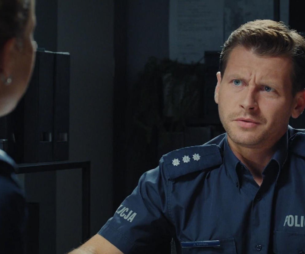 Komendant Adam Karski w niebieskim policyjnym mundurze patrzy na Natalkę. Scena z serialu M jak miłość, gdzie Rafał donosi Natalce o romansie Adama, budząc jej zaniepokojenie. Więcej o ich relacji znajdziesz na Super Seriale.