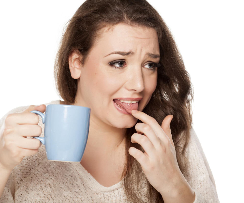 Młoda kobieta o brązowych włosach, w jasnym swetrze, trzymająca niebieski kubek, z wyraźną mimiką niezadowolenia, dotyka palcem języka, co symbolizuje odczuwanie nieprzyjemnego, słonego smaku w ustach. O przyczynach tego zjawiska przeczytasz na portalu Poradnik Zdrowie.